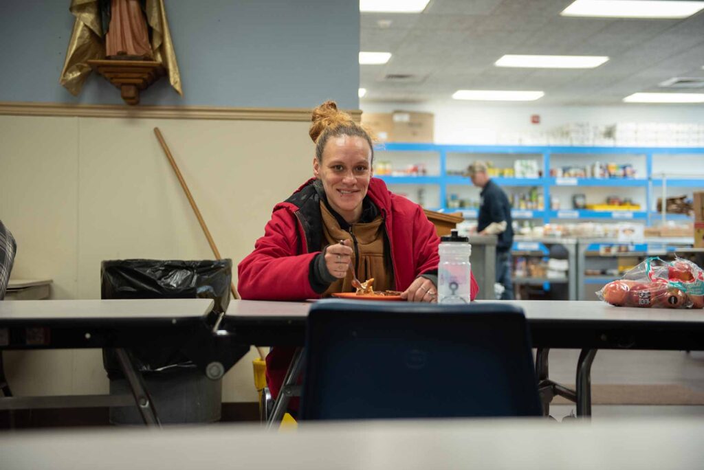 A smiling young woman in a red coat dines at a Catholic Charities facility. The space is clean and bright.