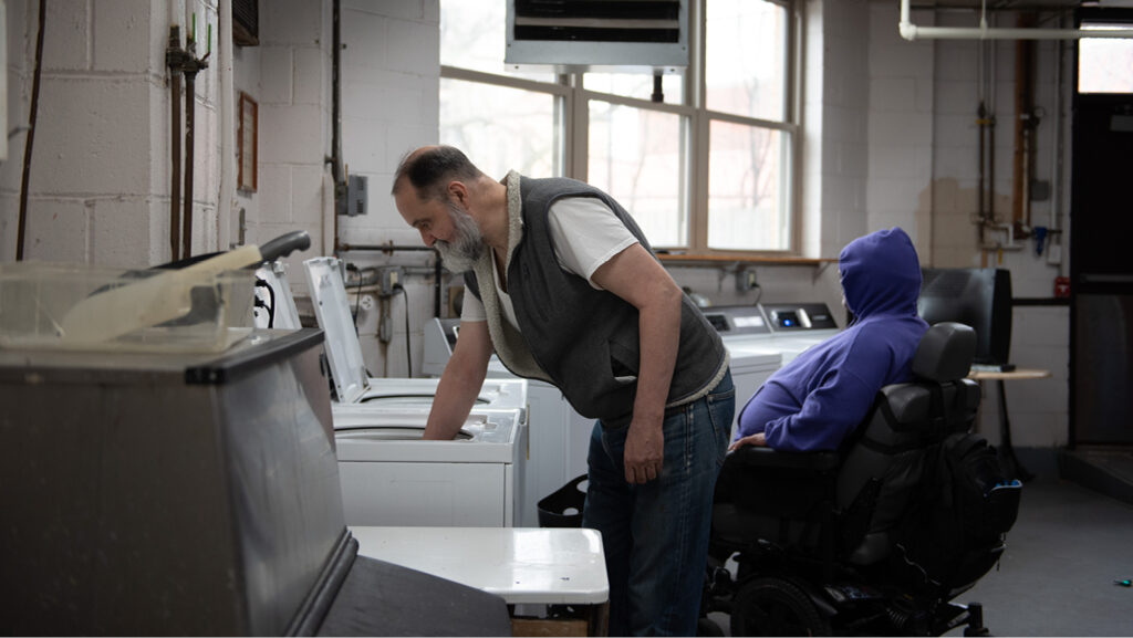 At a Catholic Charities housing site, a man with a beard in a fleece vest bends over a washing machine, checking on his clothes. Another man, wearing a purple hoodie, sits nearby.