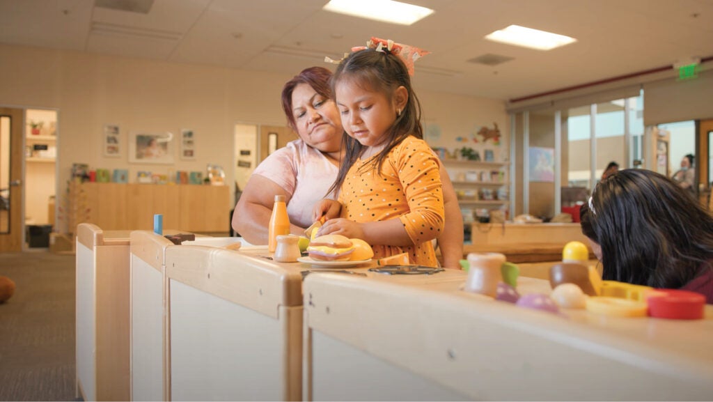 A woman watches her little girl play in a toy kitchen. The little girl is in a yellow polka dotted dress and has ribbons in her hair.