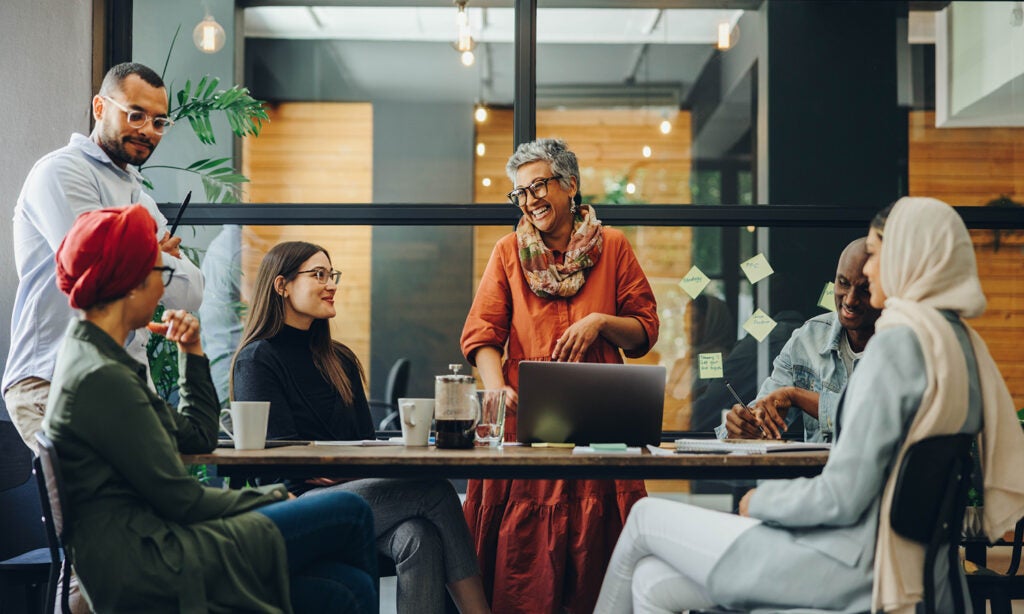 An eclectic group of people meets in an open office space. The woman leading the meeting is laughing and wearing an orange dress and scarf. There's an open laptop and a French press coffee maker on the table.