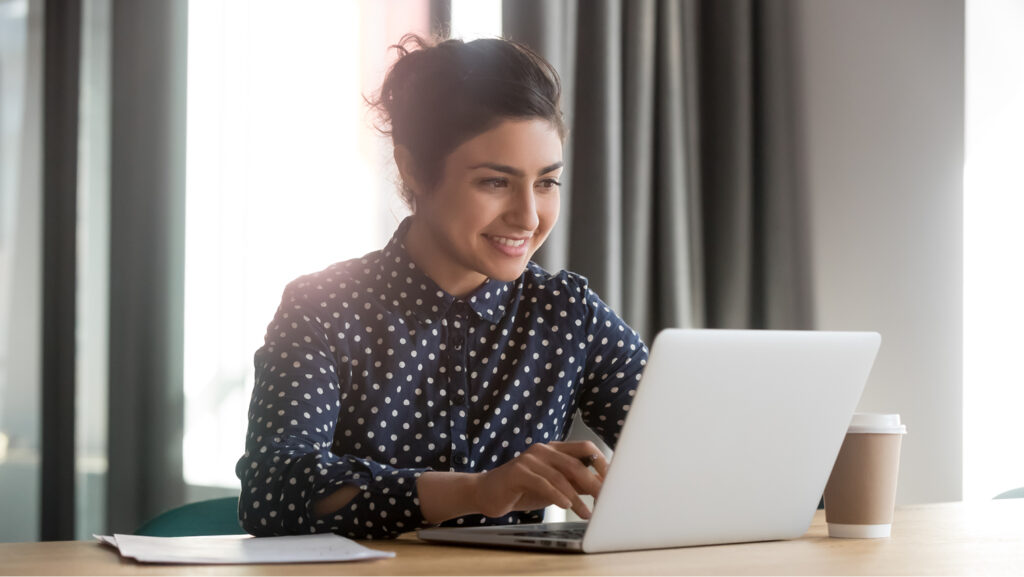 A smiling young woman with dark hair in a blue polka dot blouse works on a laptop computer. There a cup of coffee on the desk.
