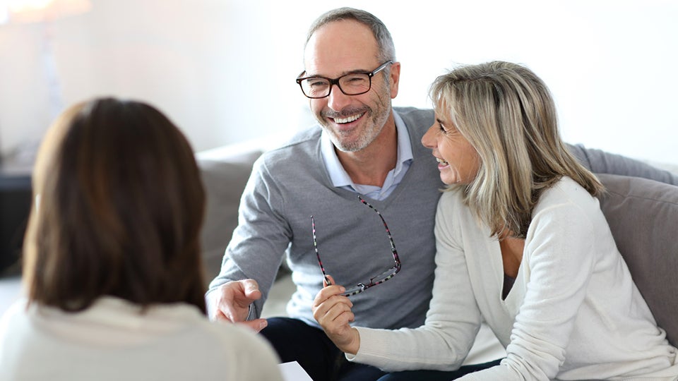 A smiling middle-aged couple sit on a couch. They are wearing v neck sweaters and she holds her glasses in her right hand. They're speaking with an investment advisor.