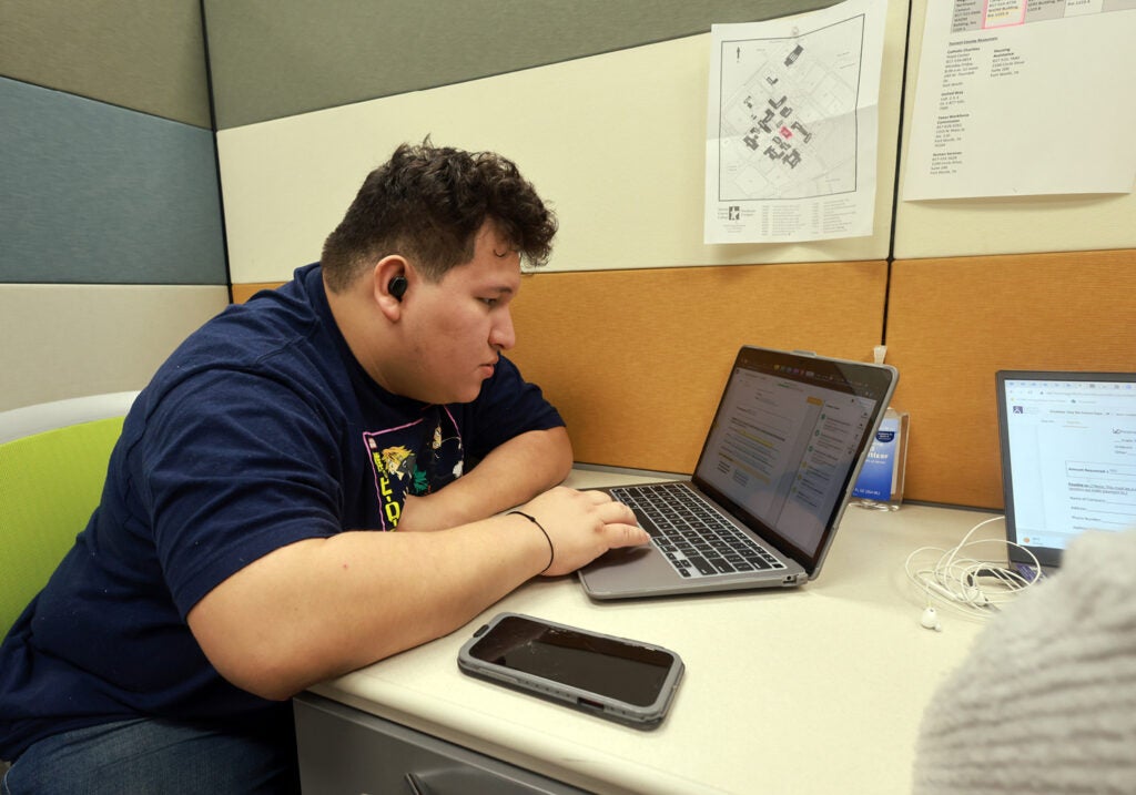 A young man in a t-shirt sits in an office space working on a laptop computer. 