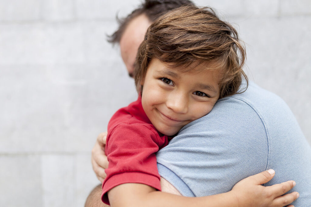 A little boy in a red shirt with brown hair smiles broadly at the camera. He is being hugged by his father and rests his head against his father's shoulder.