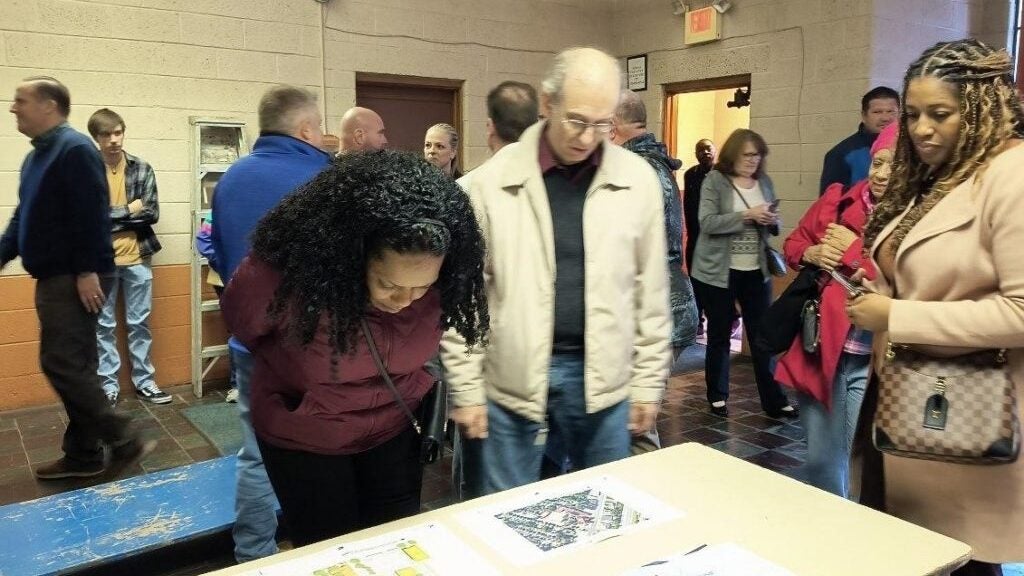 People mill around in what looks like a gym or auditorium, looking at architectural plans for a building.