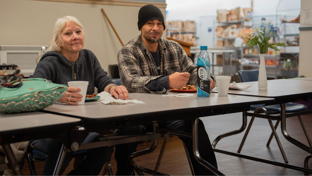 A woman and a man dine at a Catholic Charities congregate dining center. She has white hair and wears a hoodie, he has a plaid shirt on and a dark knit cap.
