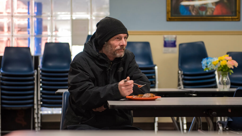 A man in a dark hoodie and knit cap sits at a dining table at a Catholic Charities congregate dining center. He has an orange plate with a full meal on it. There is a vase with yellow and blue flowers on the table behind him.