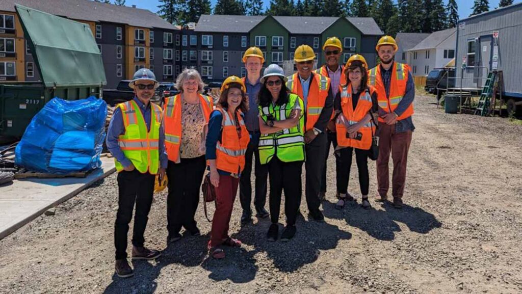 A group of smiling people in construction helmets and safety vests standing in front of a Catholic Charities affordable housing development that is under construction.