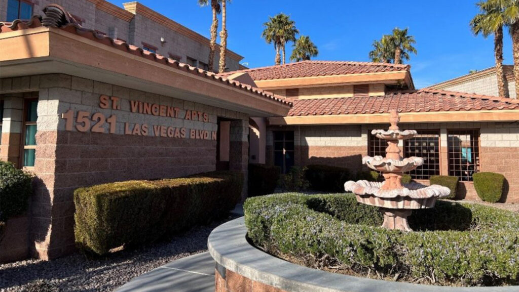 The front of the St. Vincent Apartments building, a Catholic Charities housing development in Las Vegas. The development is a low, Mediterranean style group of buildings with red tiled roofs. There is a concrete fountain in the front in the middle of a circular stand of bushes.