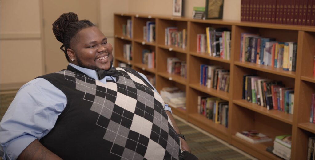 Mike, a graduate of Christopher Place Employment Academy, a program of Catholic Charities of Baltimore, is seated with a row of stocked bookshelves behind him and pictured with a big smile, wearing a blue oxford shirt, black bow tie and gray and white argyle sweater vest.