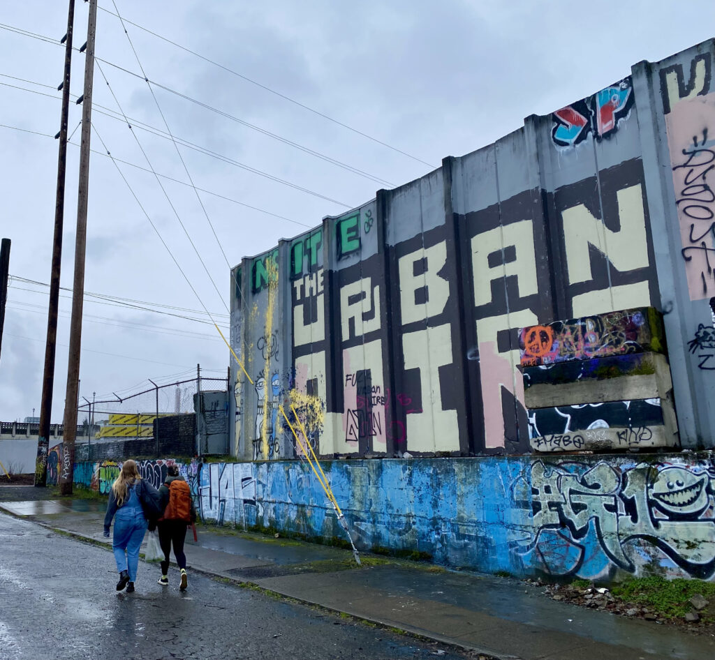 Two Catholic Charities staff members, young women with backpacks, walk with their backs to the camera alongside a tall graffitied wall. They are in an urban setting.