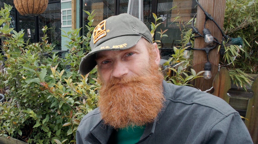 A man with a red beard and a US Army cap sits in front of shrubbery, outside a Catholic Charities center.