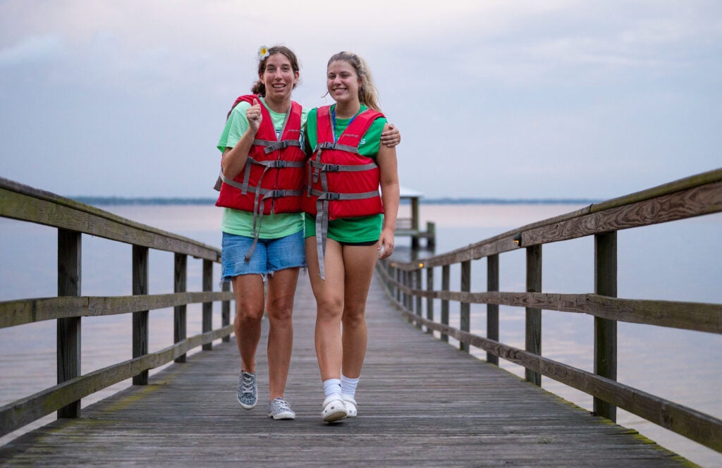 A camper and her buddy, both wearing life jackets, walk along a dock at Camp I Am Special, a program of Catholic Charities Jacksonville