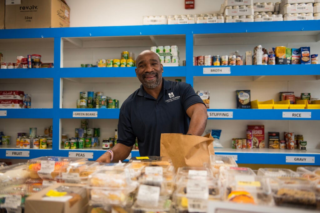 A man, wearing a navy polo and smiling broadly, packs bags of groceries in a food pantry.